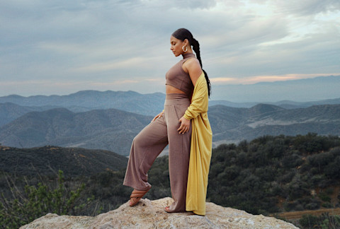 Alicia Keys standing on a mountain with a two piece outfit and a yellow sweater draping off her shoulders