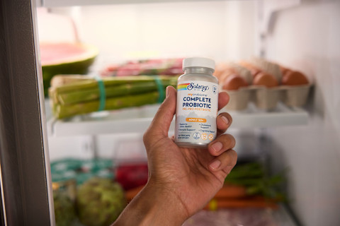 A hand holds a bottle of Solaray mycrobiome Complete Probiotic for adults over 50, reaching into a refrigerator. The inside of the fridge is visible in the background, showing shelves stocked with fresh groceries like a watermelon, asparagus, and a carton of eggs.