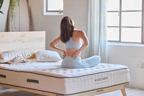 A woman wearing a light blue athletic outfit is sitting on the edge of an Avocado Extra Firm Mattress. Seen from the back, she has her hands on her lower back, as if she is stretching or in discomfort. The mattress, which has a quilted top and green piping, is on a simple, light-colored wooden bed frame. The brand name "AVOCADO" and the description "EXTRA FIRM MATTRESS" are clearly visible on the side. The setting appears to be a bright, minimalist bedroom with a large window and a white brick wall.