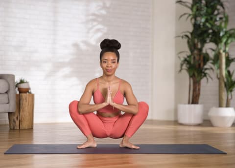 Phyllicia wearing coral yoga set doing a malasana squat on a black yoga mat