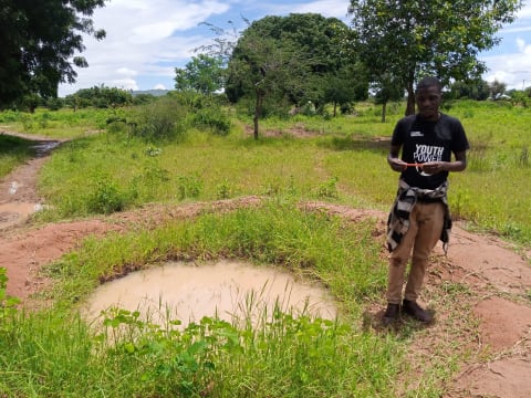 man standing next to a small, dirty pond in grassy field
