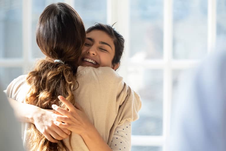 Two girls hugging, one facing the camera smiling with her eyes closed, the other has her back to the camera with a long brunette ponytail. Two girls hugging, one facing the camera smiling with her eyes closed, the other has her back to the camera with a long brunette ponytail.