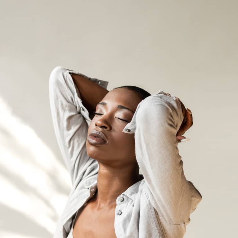 An expressive young African shaved woman stretching and holding hands above head closing her eyes. She is wearing a grey top and shirt and gold earrings.