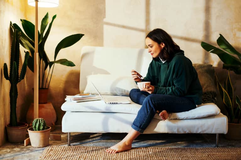 Winter shot of woman looking cozy inside in sweater on couch in living room with healthy houseplants – eating food and watching show on laptop