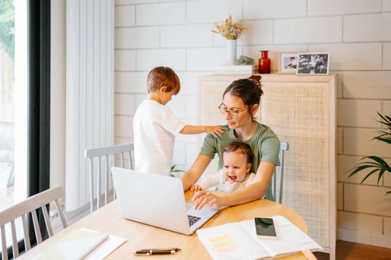 Busy concentrated mother using laptop with weepy baby on the lap near active kid on a chair at modern home