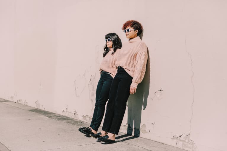 Two matching girls in pink sweaters, black jeans, and white sunglasses. These best friends are learning against a wall.