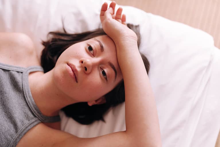 Brunette woman lying on her bed wearing a grey tank top, looking tired or distressed, resting her forearm on her forehead.