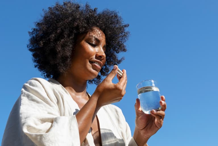 Black woman taking two multivitamin capsule holding a glass of water with a blue sky in the background