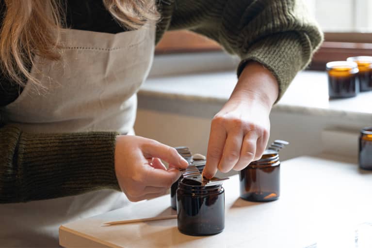 White woman wearing green sweater and tan apron adding the wick to homemade candle