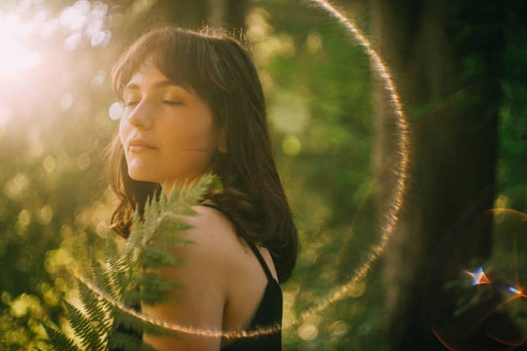 Portrait Of Young Woman With Eyes Closed And Fern In Her Hands. Standing In The Forest Catching Sun Rays