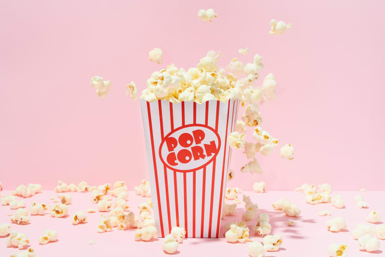 Popcorn in red striped box on pink background with kernels flying around