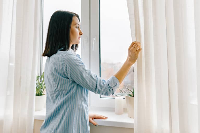 Young woman in blue shirt opening curtains looking out the window in the morning in the room, cloudy day in the city