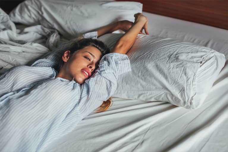 Blonde woman in a striped button down stretching in bed, sunlight beaming in.