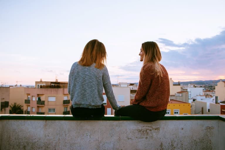 Two women sit on a wall overlooking a city at sunset