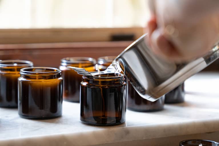 White hand holding silver sauce pan, pouring melted wax into amber jar for a homemade candle