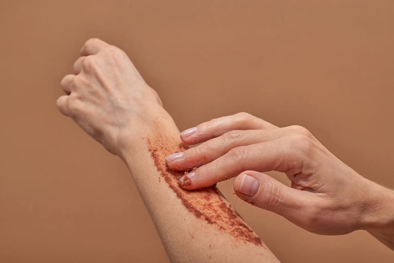 Anonymous woman smearing scrub on arm during skin care routine against brown background