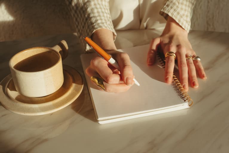 Hands in the sunlight writing in a notepad on top of marble table with a cup or coffee next to it