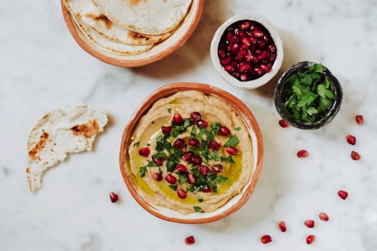 Overhead of hummus, pita bread, pomegranate arils, and fresh parsley on marble counter top